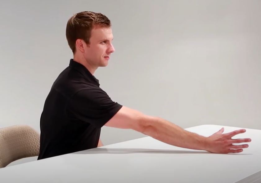 A White man is sitting at a table with his right arm outstretched in front of him grasping an imaginary object.
