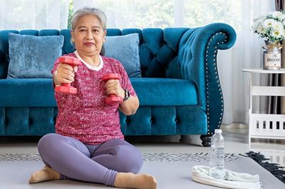 A senior Asian woman is sitting on her living room floor. She is lifting dumbbells to her chest. A senior Asian woman is sitting on her living room floor. She is lifting dumbbells to her chest.
