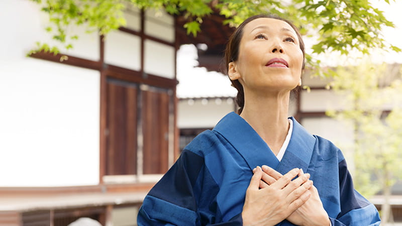 Una mujer japonesa de edad avanzada, vestida con un kimono azul, tiene las manos apoyadas sobre el pecho con un gesto de sanación mientras mira hacia el cielo.