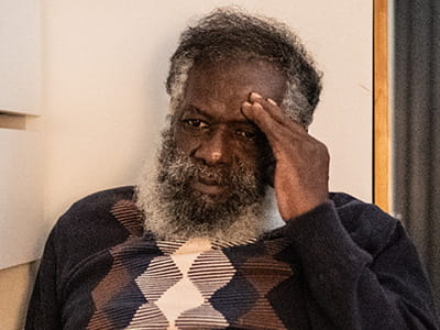 An older Somali man is sitting against a wall holding his head.