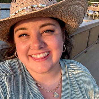 Ashleigh Mackey is smiling in a straw hat with water and a pier in the background.