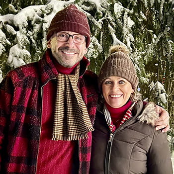 John and Melissa Rataiczak are wearing cold weather accessories and posing in front of a snowy pine tree.
