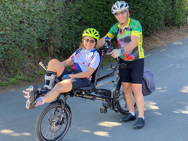 Debra Meyerson and Steve Zuckerman are dressed in cycling gear and posing with their tandem bike in an outdoor setting. Debra Meyerson and Steve Zuckerman are dressed in cycling gear and posing with their tandem bike in an outdoor setting.
