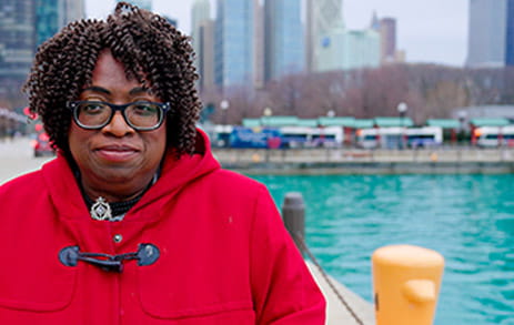 Stroke survivor, Minnie, is standing along a waterway in a red coat with city buildings in the background.