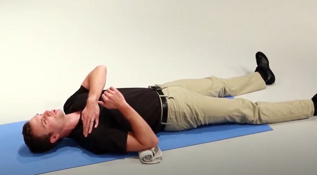 A White man is lying on his back on a mat with his legs outstretched. He has his left hand resting on his right shoulder.
