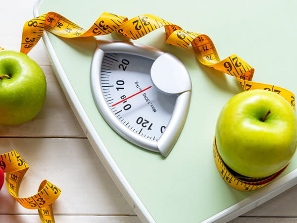 Green apples, a red dumbbell and measuring tape are arranged around a body scale.